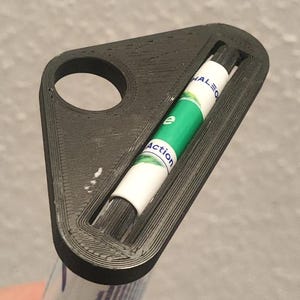May include: A black 3D-printed plastic holder with a triangular shape and a circular hole at one end. The holder features a central slot containing a white and green cylindrical inhaler with the visible text "Action" and "HALEON".