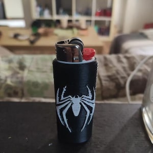 May include: A black cylindrical lighter case featuring a white spider emblem on the front. The case is made of a textured material and holds a standard metal lighter with a red ignition button and a silver-colored flint wheel mechanism.