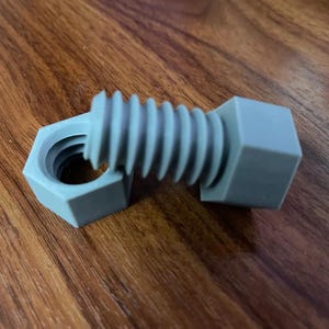 May include: A light gray plastic bolt with a hexagonal head and a matching hexagonal nut. The bolt features a threaded shaft that is partially inserted into the nut, showcasing the distinct spiral grooves and the solid, matte-finished construction.