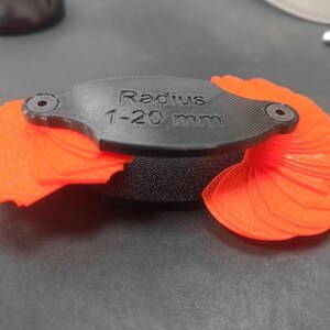 May include: A black plastic radius gauge tool with the text "Radius 1-20 mm" printed on the top. The device features two sets of bright orange fan-shaped measuring blades that extend from the central black housing on both sides.