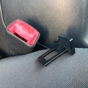 May include: A gray car seat belt buckle with a bright red release button featuring the word PRESS embossed on the surface. Next to the buckle lies a separate, small, black plastic rectangular insert piece with a hollow center and a flat tab.