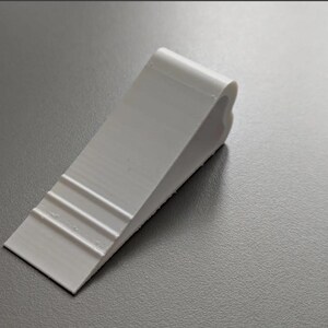 May include: A white door wedge with a slanted, tapered profile. The top surface features three horizontal ridges near the thin edge, while the thicker end has a rounded, heart-shaped contour. The object has a smooth, matte finish throughout.