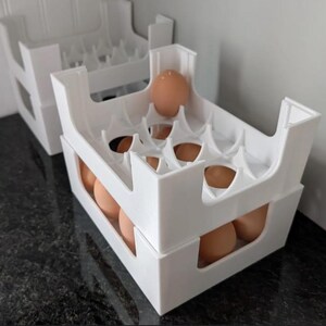 May include: A white plastic egg storage crate with a stackable design, featuring molded compartments to hold brown eggs securely. The structure has open sides and a grid-like interior, allowing for visibility and organization of multiple eggs.