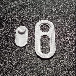 May include: Two white 3D printed plastic components. One piece is a small oval with a raised circular stud on the surface. The second piece is a larger oval frame featuring a top circular opening and a bottom elongated oval opening with textured edges.
