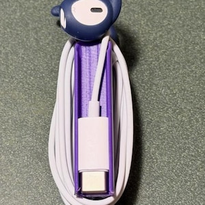 May include: A dark blue earbud with a curved hook design rests atop a purple plastic cable organizer. A white charging cable is neatly coiled around the central purple holder, with the metallic USB-C connector positioned at the bottom end.