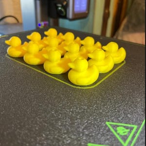 May include: A collection of small, bright yellow 3D-printed rubber duck figures arranged in a grid pattern on a dark, textured 3D printer build plate. Each duck features a rounded body, a small tail, and a distinct beak, all created in a uniform matte finish.