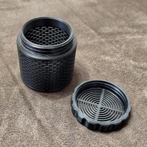 May include: A black cylindrical container with a threaded top and a matching circular lid. The body of the container features a repeating grid-like mesh pattern, while the lid has a concentric circular grate design with a cross-shaped support.