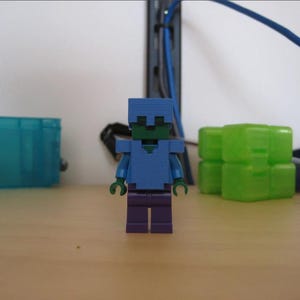 Op de afbeelding: Een kleine plastic blokfiguur met een groen hoofd en zwarte ogen, voorzien van een blauwe helm en blauwe borstplaat over een blauw bovenlichaam, met donkerpaarse benen en groene handen. De figuur heeft een blokkerig, gepixeld uiterlijk.