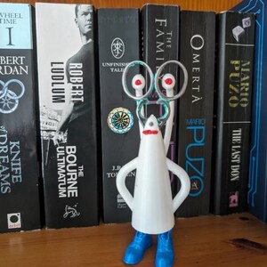 May include: A white cone-shaped figurine with blue shoes, a red mouth, and two tall stalks topped with silver rings. The figure stands upright, featuring a smooth body and arms akimbo, positioned in front of a row of various book spines on a shelf.