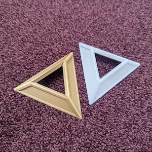May include: Two triangular frames, one in a metallic gold finish and one in a matte white finish. Each frame features a raised, beveled edge design with a hollow center, creating a geometric shape that is open in the middle for display purposes.