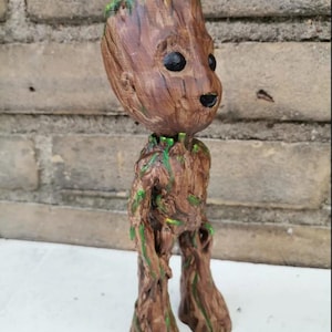May include: A brown, tree-like humanoid figurine with a textured bark surface, large black eyes, and small patches of bright green paint accents on the head, shoulders, and torso. The figure stands upright on two legs with a slender, branch-like body.