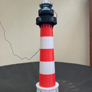 Könnte beinhalten: Ein Leuchtturmmodell mit einem zylindrischen Turm mit abwechselnden horizontalen roten und weißen Streifen. Es hat einen weißen runden Sockel mit Bogenöffnungen, ein schwarzes Geländer und einen oberen Teil mit Licht und roter Spitze.
