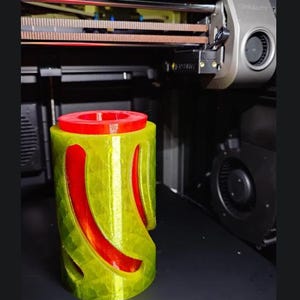 Könnte beinhalten: Ein 3D-gedrucktes zylindrisches Objekt mit einem leuchtend roten Innenkern und einer durchscheinenden gelben Außenhülle mit geschwungenen vertikalen Ausschnitten. Das Objekt steht auf dem Druckbett eines Creality 3D-Druckers mit sichtbaren mechanischen Teilen.