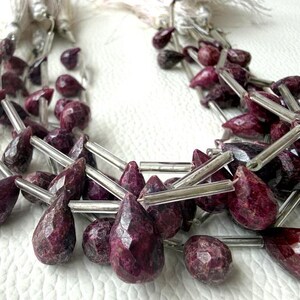 May include: A multi-strand necklace featuring faceted, teardrop-shaped dark magenta gemstone beads interspersed with clear, cylindrical glass spacer beads. The necklace is finished with white tassels at the ends of the strands.
