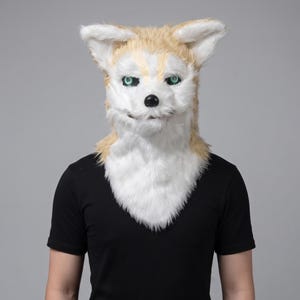 Puede incluir: Una máscara de animal de cabeza completa con piel sintética realista en tonos beige y blanco. La máscara tiene orejas puntiagudas, ojos de color verde brillante y una nariz negra. El pelaje texturizado cubre la cabeza y el cuello.