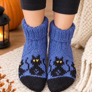 Puede incluir: Calcetines tobilleros de punto azul con un patrón de gatos negros de ojos amarillos y punteras negras sólidas. Los calcetines tienen un puño grueso acanalado en la parte superior y están confeccionados con una textura de punto grueso.