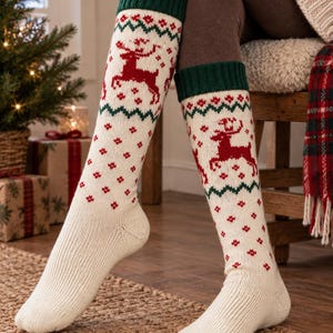 May include: Knee-high knit socks featuring a cream base with a red reindeer pattern, green ribbed cuffs, and a small red geometric motif. The socks have a ribbed texture on the foot and a fair isle style design on the calf with green zig-zag borders.