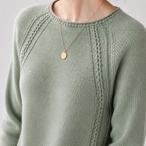 May include: A sage green long-sleeved knit sweater featuring a round neckline and raglan sleeves with vertical braided cable knit detailing along the shoulder seams, paired with a thin gold chain necklace holding a small round pendant.