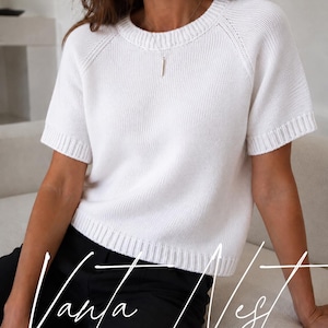 Könnte beinhalten: Ein kurzärmeliger, weißer Strickpullover mit Rundhalsausschnitt und gerippten Abschlüssen an Kragen, Ärmeln und Saum. Das Kleidungsstück hat ein schlichtes, einfarbiges Design mit glatter Textur. Der Text "Vanja Nest" ist in weißer Schrift unten zu sehen.
