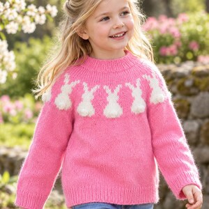 May include: A bright pink long-sleeved knit sweater featuring a crew neckline and a repeating pattern of white knitted rabbits across the chest. The sweater has a ribbed collar, cuffs, and hem, with textured white pom-poms forming the rabbit tails.