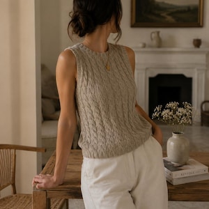 Può includere: Un gilet smanicato in maglia beige con motivo a trecce verticali, scollo rotondo e orlo a coste. Il capo è realizzato con un filato dalla finitura testurizzata ed è indossato con pantaloni bianchi a tinta unita.
