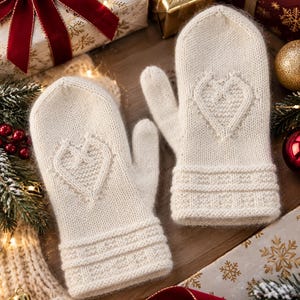 Könnte beinhalten: Ein Paar cremefarbene Strickfäustlinge mit einem strukturierten herzförmigen Design auf dem Handrücken und einem gerippten Bündchen mit horizontalem Gittermuster. Die Fäustlinge haben eine weiche, flauschige Textur sowie abgerundete Daumen.