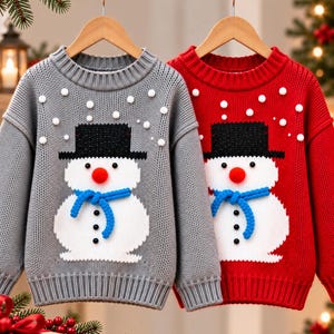 Könnte beinhalten: Zwei langärmelige Strickpullover, einer in Grau und einer in Rot, jeweils mit einem Schneemann-Motiv. Der Schneemann hat einen schwarzen Zylinder, eine orangefarbene Nase, einen blauen Schal und schwarze Knöpfe. Kleine weiße Bommeln sind auf der Brust verteilt.