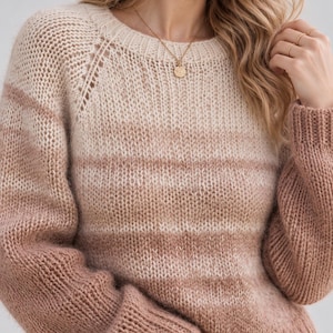 Pode incluir: Uma camisola de malha de manga comprida com um padrão de riscas horizontais em tons de creme, bege e rosa seco. A peça apresenta um decote redondo, punhos canelados e uma construção em malha texturada com detalhes visíveis nas costuras.