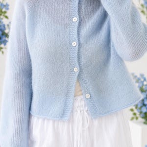 Puede incluir: Un cárdigan de punto azul claro, difuso y de manga larga, con cuello redondo y cierre frontal de botones con cinco botones blancos pequeños y redondos. La prenda tiene un aspecto suave y texturizado, y se combina con pantalones blancos.