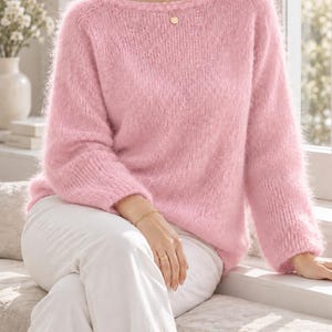 May include: A soft, fuzzy, light pink long-sleeved jumper with a wide boat neckline. The knit garment features a textured, brushed surface and ribbed cuffs at the wrists. The jumper is paired with plain white trousers, creating a simple, clean look.
