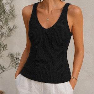 Könnte beinhalten: Ein schwarzes ärmelloses Tanktop mit tiefem V-Ausschnitt und vertikaler Rippstrickstruktur. Das Kleidungsstück ist körpernah geschnitten, hat breite Schulterträger und einen geraden Saum auf Taillenhöhe und besteht aus einfarbigem Strickstoff.