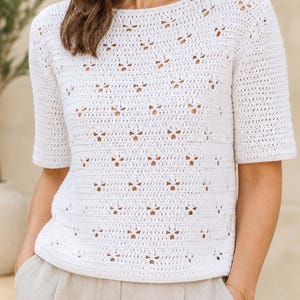 May include: A white short-sleeved top featuring an intricate crochet pattern with repeating openwork flower-like motifs. The garment has a round neckline and a straight hem, crafted with a consistent stitch throughout the body and sleeves.