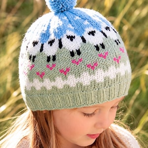 Puede incluir: Un gorro de punto con un pompón azul claro en la parte superior. El gorro presenta un patrón repetitivo de ovejas blancas con detalles negros sobre un fondo verde salvia, con acentos rosas y una franja blanca en zigzag.