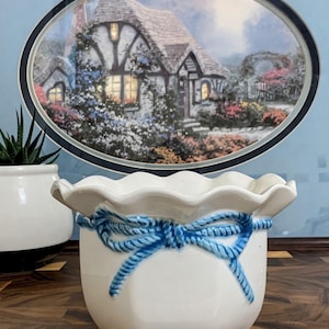 May include: A white ceramic planter with a scalloped rim and a raised, blue painted rope-style bow detail wrapped around the upper exterior. The planter has a smooth, glossy finish and a hexagonal base shape, providing a clean and simple aesthetic.