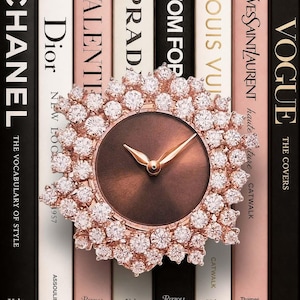 Può includere: Un orologio analogico rotondo con quadrante marrone scuro e lancette in oro rosa, circondato da una cornice circolare di numerose pietre chiare e scintillanti. L'orologio è posizionato davanti a una fila di libri verticali con dorsi colorati.