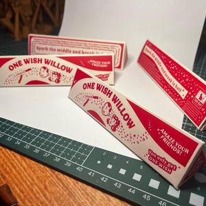 May include: Three triangular cardboard boxes with red and white graphics. The text reads ONE WISH WILLOW, AMAZE YOUR FRIENDS, and You only get ONE WISH. Each box features a cartoon illustration of two faces and stars, with instructions to spark the middle and break in half.