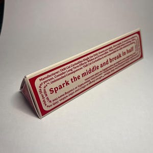 May include: A triangular prism-shaped object with a white surface and a red border. The text reads: Spark the middle and break in half. Manufacturer: TABI Cat Curiosities Single Use Only: Grants one wish. Once made, it cannot be undone.
