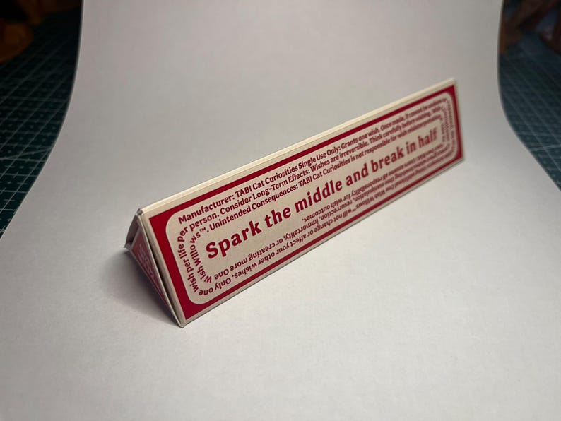 May include: A triangular prism-shaped object with a white surface and a red border. The text reads: Spark the middle and break in half. Manufacturer: TABI Cat Curiosities Single Use Only: Grants one wish. Once made, it cannot be undone.