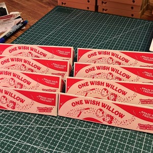 May include: A collection of rectangular, cream-colored paper boxes with red text and graphics. Each box features the text "ONE WISH WILLOW," "AMAZE YOUR FRIENDS!," and "You only get ONE WISH," along with illustrations of two faces and a small wand.