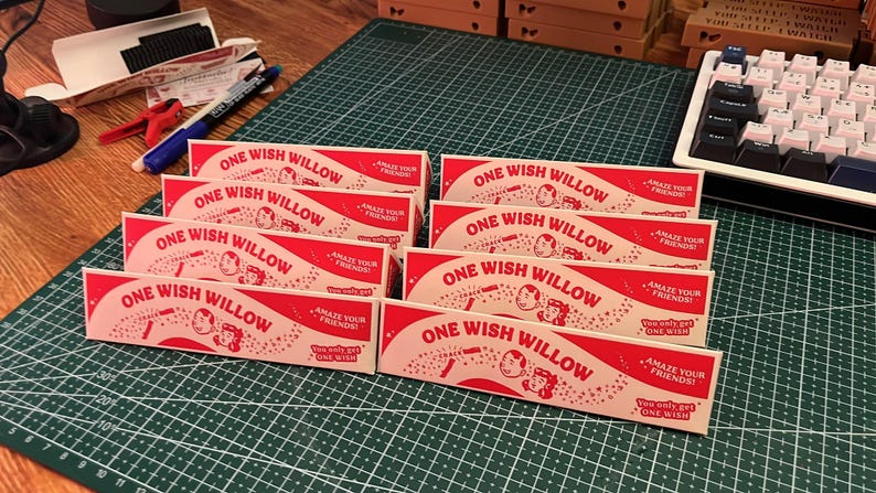 May include: A collection of rectangular, cream-colored paper boxes with red text and graphics. Each box features the text "ONE WISH WILLOW," "AMAZE YOUR FRIENDS!," and "You only get ONE WISH," along with illustrations of two faces and a small wand.