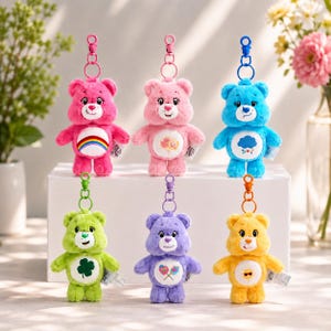 May include: A set of six plush bear keychains in various colors including pink, light pink, blue, green, purple, and yellow. Each bear features a unique belly badge, a plastic swivel clasp, and a white circular patch on the chest with a graphic.