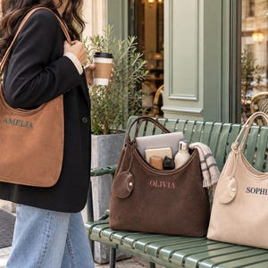 Puede incluir: Tres bolsos tipo hobo de textura suave en color canela, marrón oscuro y beige. Cada bolso cuenta con un dije circular, dos correas para el hombro y un nombre bordado en el frente: Amelia, Olivia y Sophia. Diseño de apertura superior.