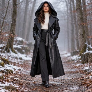 Puede incluir: Una gabardina larga de cuero negro con una capucha grande, usada sobre un suéter de cuello alto color crema y pantalones negros. El abrigo cuenta con una cintura ceñida con cinturón, mangas largas con puños y un acabado liso y elegante.