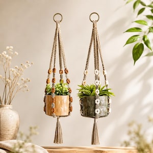 May include: Two macrame plant hangers made of beige cord, each featuring a metal ring at the top and a tassel at the bottom. One hanger holds a light orange ceramic pot with brown beads, and the other holds a dark green pot with white beads.