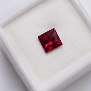 May include: A square-cut, deep red gemstone with faceted surfaces, displayed centered within a white, square-shaped presentation box. The stone exhibits a vibrant, saturated crimson hue and sharp, clean edges, resting on a soft white foam insert.