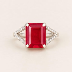 Op de afbeelding: Een zilverkleurige ring met een grote, rechthoekige, smaragdgeslepen rode edelsteen vastgehouden door vier klauwen. De gespleten ringband is versierd met kleine, ronde, heldere pavé-gezette steentjes die vanaf het midden doorlopen.