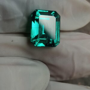 May include: A vibrant emerald green gemstone with a rectangular emerald cut, featuring sharp facets and internal reflections. The stone displays a deep, saturated green hue with visible internal inclusions and a polished, reflective surface.