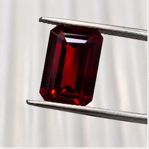 Può includere: Una gemma rosso scuro con taglio a smeraldo tenuta da una pinzetta metallica color argento. La pietra presenta una superficie sfaccettata con bordi netti e una forma rettangolare, mostrando una ricca tonalità cremisi traslucida.