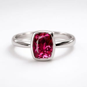 Puede incluir: Un anillo de tono plateado con una piedra preciosa de color rosa intenso en talla cojín, engastada en un bisel liso. La banda es pulida y metálica, mostrando una superficie limpia y reflectante que resalta la piedra facetada central.