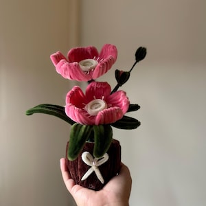Può includere: Una composizione floreale artigianale con due fiori dalla consistenza vellutata in rosa acceso con centri bianchi. I fiori presentano foglie e steli in velluto verde scuro, inseriti in un vaso marrone testurizzato con un fiocco bianco.